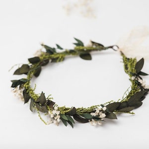 May include: A handmade floral crown with dark green leaves, small white flowers, and sprigs of greenery. The crown is tied with a cream ribbon. This natural headpiece is suitable for weddings or special occasions.