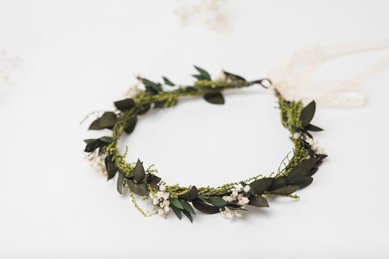 May include: A handmade floral crown with dark green leaves, small white flowers, and sprigs of greenery. The crown is tied with a cream ribbon. This natural headpiece is suitable for weddings or special occasions.