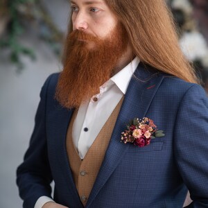 May include: A man wearing a blue suit jacket with a brown vest and a white shirt. He has long red hair and a red beard. He is wearing a small bouquet of flowers on his lapel.