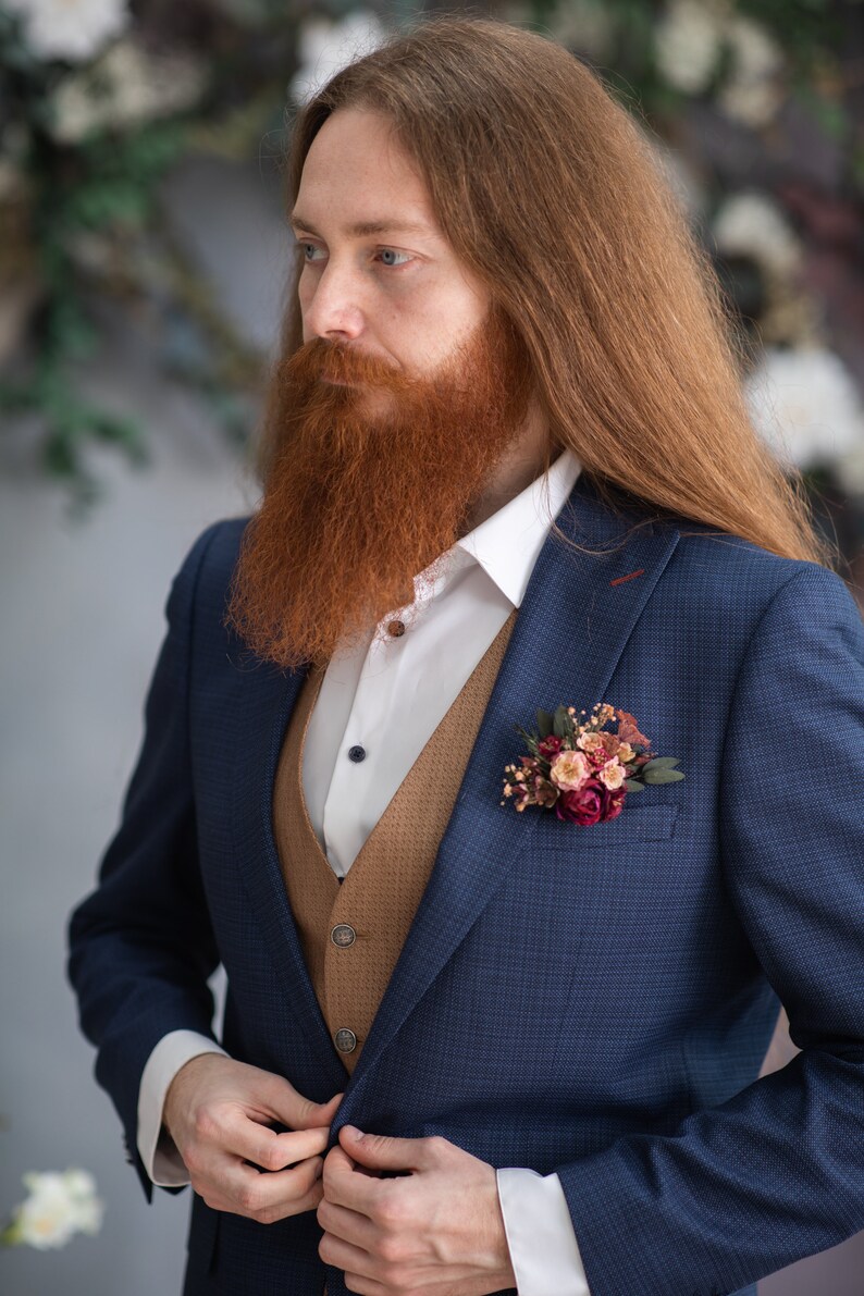 May include: A man wearing a blue suit jacket with a brown vest and a white shirt. He has long red hair and a red beard. He is wearing a small bouquet of flowers on his lapel.