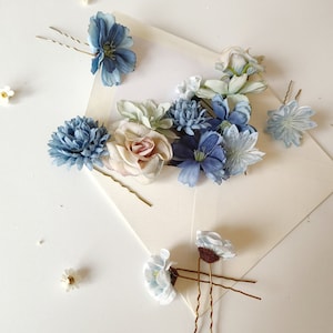 K&ouml;nnte beinhalten: Eine Auswahl an floralen Haarnadeln in Blau-, Wei&szlig;- und Rosat&ouml;nen. Die Nadeln weisen verschiedene Blumendesigns, darunter Rosen und G&auml;nsebl&uuml;mchen, auf und sind auf einer wei&szlig;en Oberfl&auml;che angeordnet. Haarschmuck f&uuml;r Hochzeiten oder besondere Anl&auml;sse.