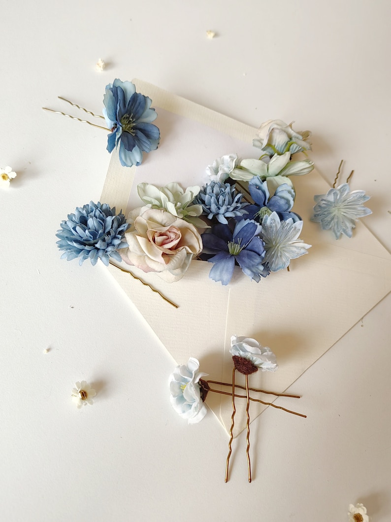 K&ouml;nnte beinhalten: Eine Auswahl an floralen Haarnadeln in Blau-, Wei&szlig;- und Rosat&ouml;nen. Die Nadeln weisen verschiedene Blumendesigns, darunter Rosen und G&auml;nsebl&uuml;mchen, auf und sind auf einer wei&szlig;en Oberfl&auml;che angeordnet. Haarschmuck f&uuml;r Hochzeiten oder besondere Anl&auml;sse.