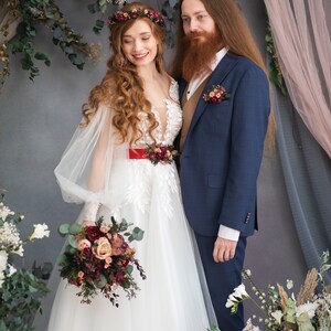 May include: A bride and groom pose for a wedding portrait. The bride is wearing a white gown with a red sash and a floral crown. The groom is wearing a blue suit with a brown vest and a floral boutonniere. They are standing in front of a floral backdrop.