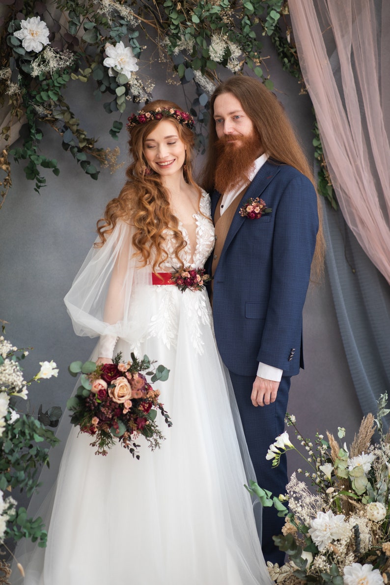 May include: A bride and groom pose for a wedding portrait. The bride is wearing a white gown with a red sash and a floral crown. The groom is wearing a blue suit with a brown vest and a floral boutonniere. They are standing in front of a floral backdrop.