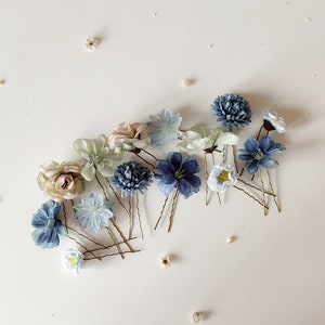 K&ouml;nnte beinhalten: Eine Auswahl an floralen Haarnadeln in Blau-, Wei&szlig;- und Beiget&ouml;nen. Die Haarnadeln zeigen verschiedene Blumendesigns, darunter Rosen und Chrysanthemen, mit goldfarbenen Drahtstielen. Kleine wei&szlig;e Bl&uuml;ten sind ebenfalls verstreut.