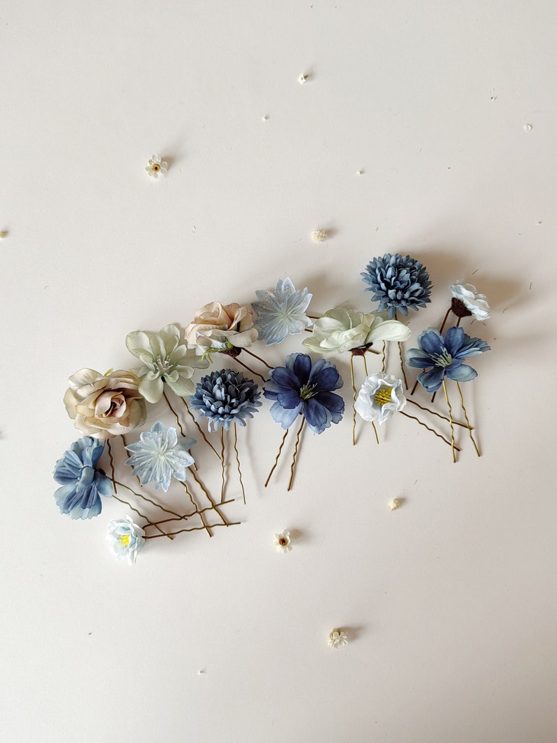K&ouml;nnte beinhalten: Eine Auswahl an floralen Haarnadeln in Blau-, Wei&szlig;- und Beiget&ouml;nen. Die Haarnadeln zeigen verschiedene Blumendesigns, darunter Rosen und Chrysanthemen, mit goldfarbenen Drahtstielen. Kleine wei&szlig;e Bl&uuml;ten sind ebenfalls verstreut.