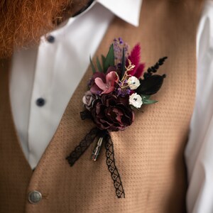 May include: A brown waistcoat with a white shirt and a floral buttonhole with burgundy, pink, and white flowers, black feathers, and a black lace ribbon.