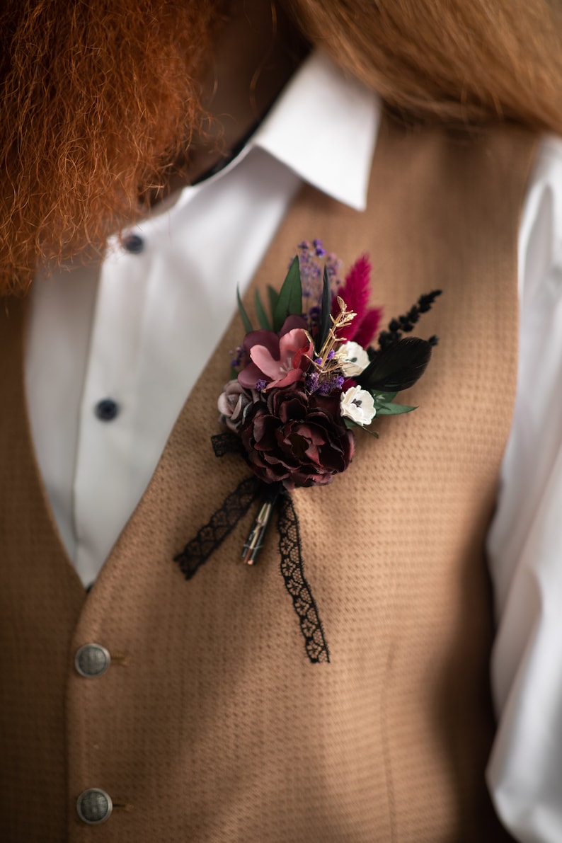 May include: A brown waistcoat with a white shirt and a floral buttonhole with burgundy, pink, and white flowers, black feathers, and a black lace ribbon.