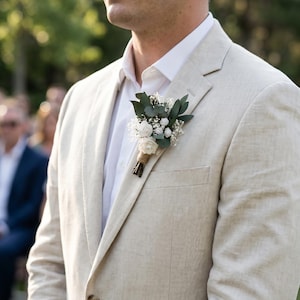 Puede incluir: Americana de lino beige claro con camisa blanca y boutonniere floral. La boutonniere presenta rosas blancas, vegetaci&oacute;n y paniculata. La americana tiene solapas con muescas y cierre de un solo bot&oacute;n.