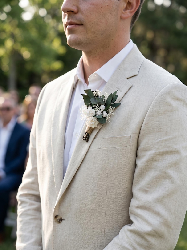 Puede incluir: Americana de lino beige claro con camisa blanca y boutonniere floral. La boutonniere presenta rosas blancas, vegetaci&oacute;n y paniculata. La americana tiene solapas con muescas y cierre de un solo bot&oacute;n.