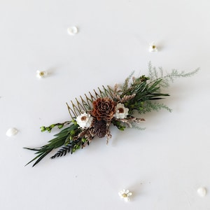 May include: A gold hair comb with a floral arrangement of dried white flowers, brown pine cones, and green foliage.