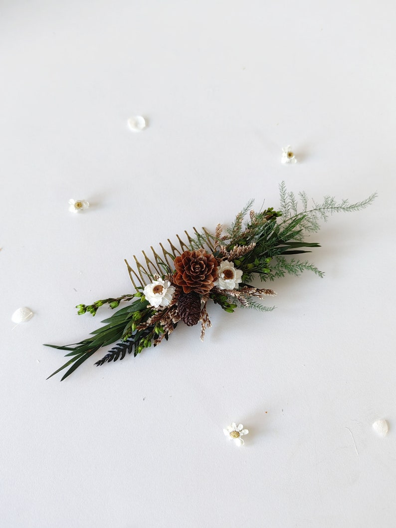 May include: A gold hair comb with a floral arrangement of dried white flowers, brown pine cones, and green foliage.