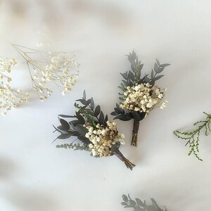 May include: Two small boutonnieres, each featuring dried white flowers and dark green leaves, bound with brown twine. Sprigs of white flowers and greenery are arranged on a white surface, creating a natural, rustic look.