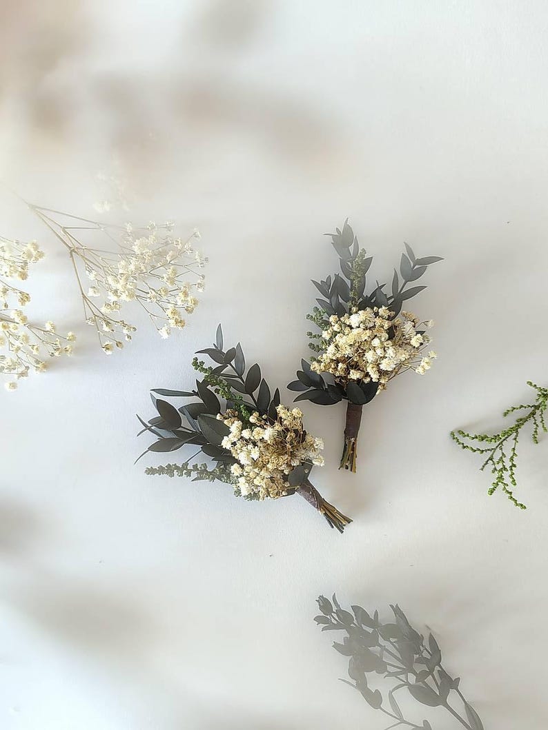 May include: Two small boutonnieres, each featuring dried white flowers and dark green leaves, bound with brown twine. Sprigs of white flowers and greenery are arranged on a white surface, creating a natural, rustic look.