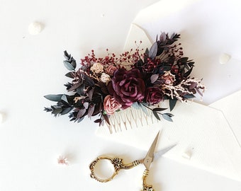 Puede incluir: Una peineta floral con rosas burdeos, hojas oscuras y pequeñas flores rosas. Junto a ella, unas tijeras doradas con detalles florales. Los artículos están sobre una superficie blanca, con un sobre blanco en el fondo.