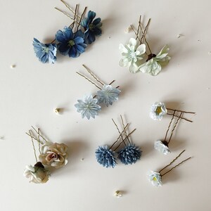 K&ouml;nnte beinhalten: Eine Auswahl an floralen Haarnadeln in verschiedenen Farben, darunter Blau, Wei&szlig; und Beige. Die Nadeln zeigen verschiedene Blumendesigns, wie Rosen und G&auml;nsebl&uuml;mchen, auf einer wei&szlig;en Oberfl&auml;che. Haarschmuck f&uuml;r Hochzeiten oder besondere Anl&auml;sse.