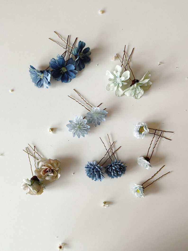 K&ouml;nnte beinhalten: Eine Auswahl an floralen Haarnadeln in verschiedenen Farben, darunter Blau, Wei&szlig; und Beige. Die Nadeln zeigen verschiedene Blumendesigns, wie Rosen und G&auml;nsebl&uuml;mchen, auf einer wei&szlig;en Oberfl&auml;che. Haarschmuck f&uuml;r Hochzeiten oder besondere Anl&auml;sse.