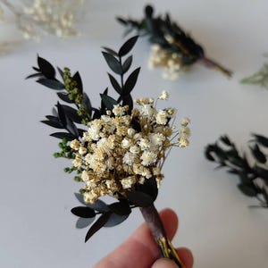 May include: A handcrafted boutonniere with dried white baby's breath flowers, dark green leaves, and black leaves. The stems are wrapped in brown floral tape. Other boutonnieres are visible, suggesting a wedding or special event theme.