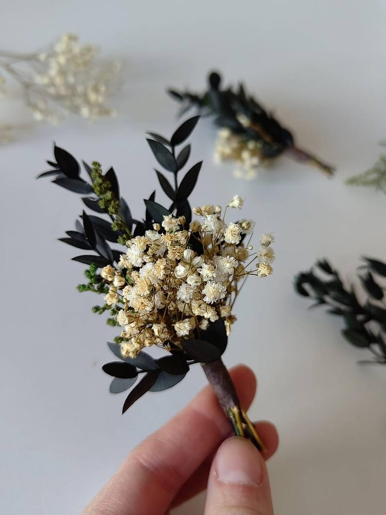 May include: A handcrafted boutonniere with dried white baby's breath flowers, dark green leaves, and black leaves. The stems are wrapped in brown floral tape. Other boutonnieres are visible, suggesting a wedding or special event theme.