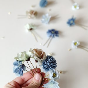 K&ouml;nnte beinhalten: Blumen-Haarnadeln in verschiedenen Blau-, Gr&uuml;n- und Beiget&ouml;nen. Die Haarnadeln haben Stoffblumen und sind auf goldfarbenen Metall-Haarnadeln angebracht. Der Hintergrund ist eine weiche, neutrale Farbe.