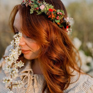 May include: A woman with long, curly red hair wears a floral crown made of dried flowers, berries, and pine cones. She is holding a sprig of white blossoms and smelling them.