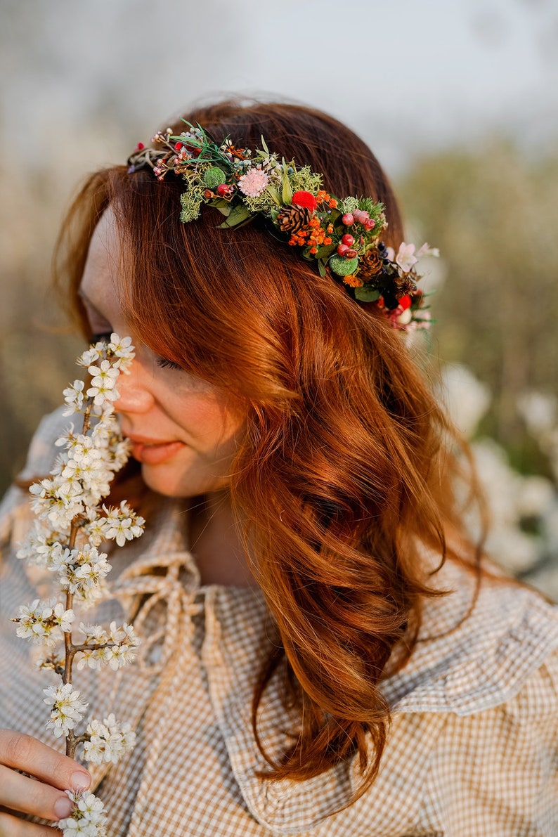 May include: A woman with long, curly red hair wears a floral crown made of dried flowers, berries, and pine cones. She is holding a sprig of white blossoms and smelling them.