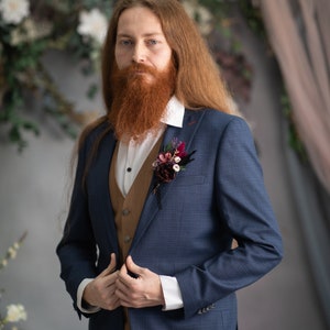 May include: A man wearing a blue suit with a brown waistcoat and a white shirt. He has long red hair and a red beard. He is wearing a small bouquet of flowers on his lapel.