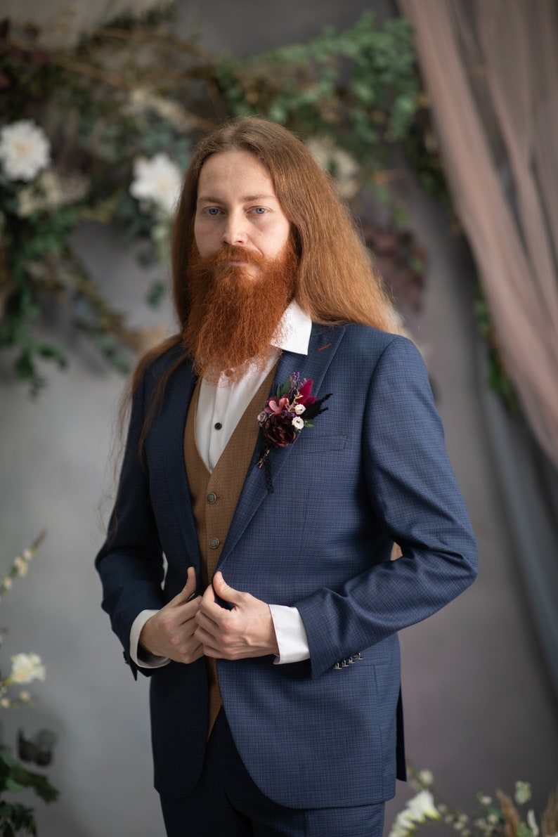 May include: A man wearing a blue suit with a brown waistcoat and a white shirt. He has long red hair and a red beard. He is wearing a small bouquet of flowers on his lapel.