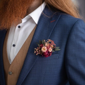 May include: A man wearing a blue suit jacket with a brown vest and a white shirt. He has a long red beard and is wearing a small bouquet of flowers in his lapel.