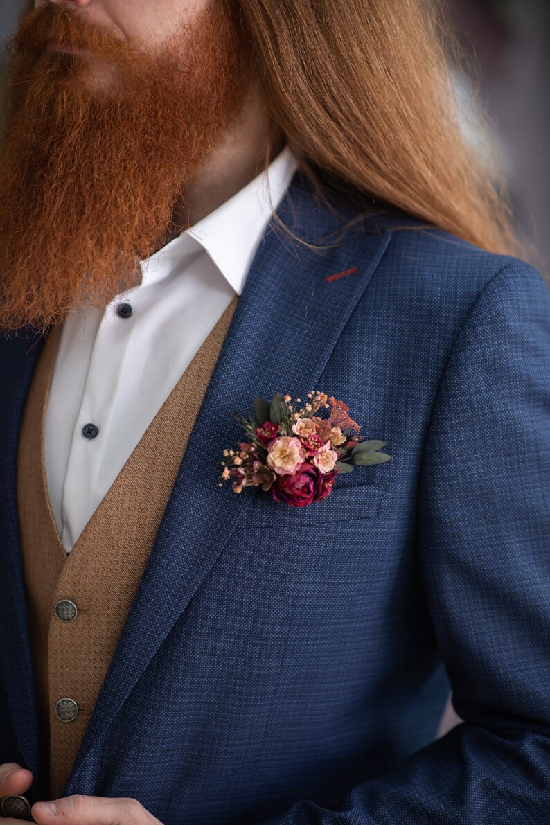 May include: A man wearing a blue suit jacket with a brown vest and a white shirt. He has a long red beard and is wearing a small bouquet of flowers in his lapel.