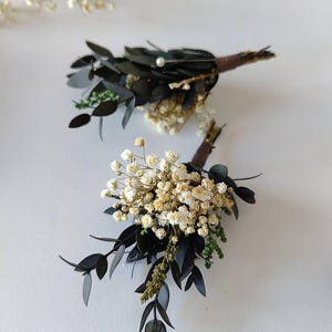 May include: Two boutonnieres featuring dark green leaves, small white flowers, and brown stems. One boutonniere has a small pearl. The boutonnieres are displayed on a white surface, with a sprig of small white flowers in the background.