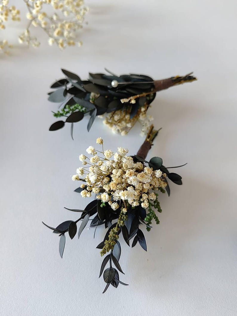 May include: Two boutonnieres featuring dark green leaves, small white flowers, and brown stems. One boutonniere has a small pearl. The boutonnieres are displayed on a white surface, with a sprig of small white flowers in the background.