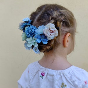 K&ouml;nnte beinhalten: Geflochtene Frisur eines Kindes, geschm&uuml;ckt mit einem Blumenkranz. Der Kranz zeigt blaue und hellrosa Bl&uuml;ten. Die Person tr&auml;gt ein wei&szlig;es, vertikal gestreiftes Hemd mit gestickten floralen Details.