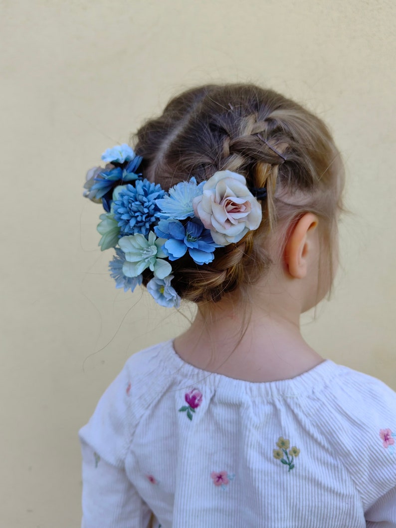 K&ouml;nnte beinhalten: Geflochtene Frisur eines Kindes, geschm&uuml;ckt mit einem Blumenkranz. Der Kranz zeigt blaue und hellrosa Bl&uuml;ten. Die Person tr&auml;gt ein wei&szlig;es, vertikal gestreiftes Hemd mit gestickten floralen Details.