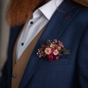 May include: A blue suit jacket with a brown vest underneath. A small bouquet of pink and burgundy flowers is pinned to the lapel.
