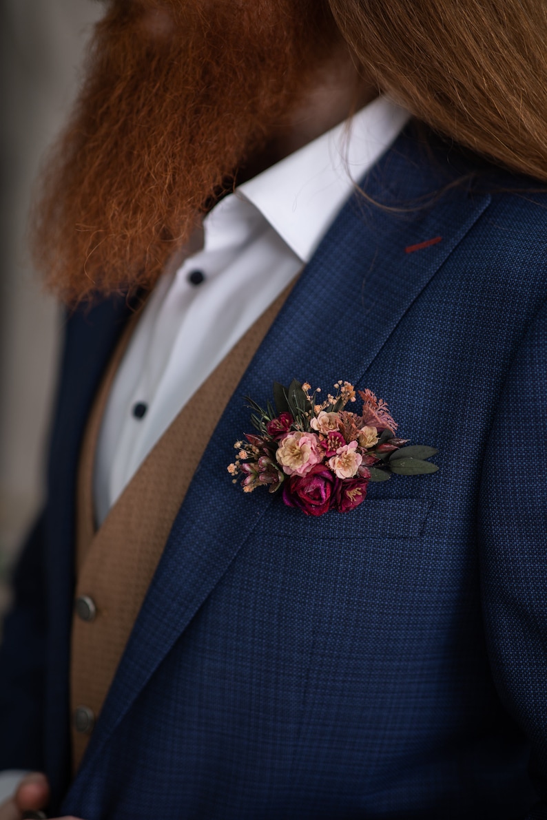 May include: A blue suit jacket with a brown vest underneath. A small bouquet of pink and burgundy flowers is pinned to the lapel.