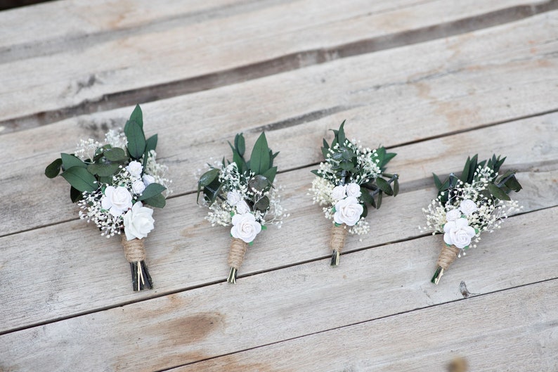 Puede incluir: Cuatro boutonnieres con acentos de rosas blancas, paniculata y hojas verde oscuro, envueltas con cuerda. Los arreglos florales se colocan sobre un fondo de madera r&uacute;stica, ideales para bodas o eventos especiales.