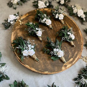 Puede incluir: Colecci&oacute;n de boutonnieres con rosas blancas, follaje verde y paniculata. Los boutonnieres est&aacute;n dispuestos sobre una rodaja de madera y una superficie gris. Cada boutonniere est&aacute; envuelto con hilo.