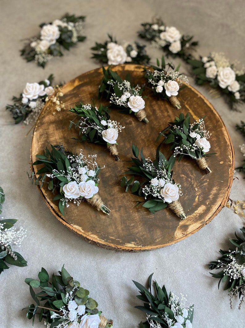 Puede incluir: Colecci&oacute;n de boutonnieres con rosas blancas, follaje verde y paniculata. Los boutonnieres est&aacute;n dispuestos sobre una rodaja de madera y una superficie gris. Cada boutonniere est&aacute; envuelto con hilo.