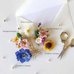 May include: Four floral hairpins with different flower arrangements. The hairpins are arranged on a white surface. The hairpins include a daisy, a sunflower, a cornflower, and a mixed arrangement of small flowers.