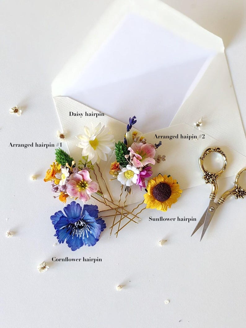 May include: Four floral hairpins with different flower arrangements. The hairpins are arranged on a white surface. The hairpins include a daisy, a sunflower, a cornflower, and a mixed arrangement of small flowers.