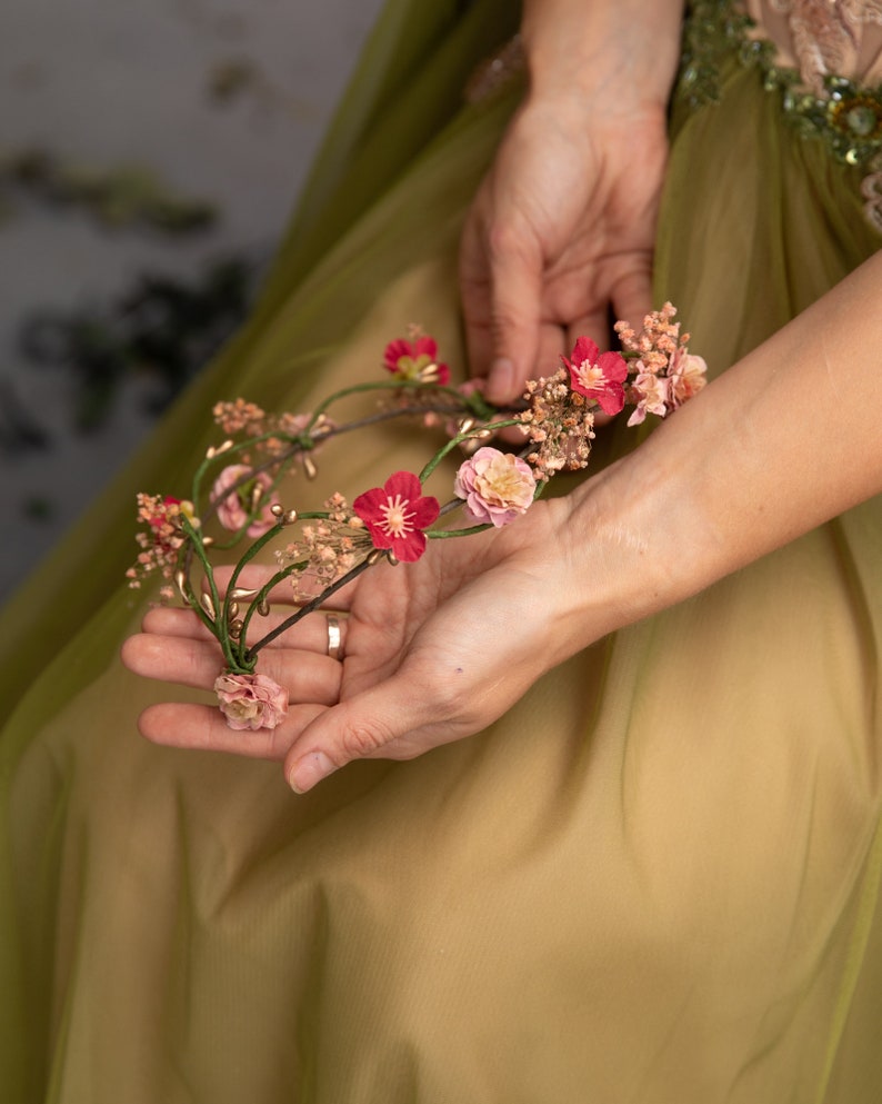 May include: A delicate floral crown with pink and red flowers, woven with thin wire and small green leaves.