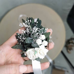 Puede incluir: Un ramillete de mu&ntilde;eca con rosas blancas, peque&ntilde;as flores blancas y hojas de color verde oscuro. Se adjunta una cinta de raso blanca para asegurar el ramillete a la mu&ntilde;eca. El arreglo floral se sostiene en una mano.