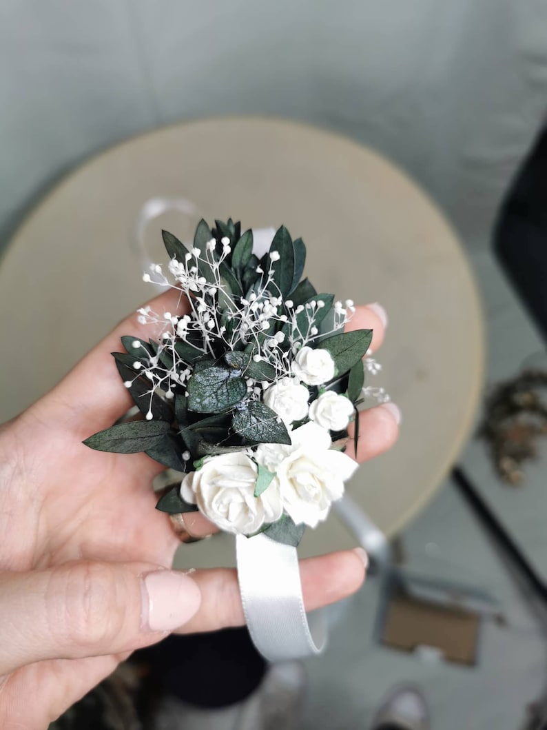 Puede incluir: Un ramillete de mu&ntilde;eca con rosas blancas, peque&ntilde;as flores blancas y hojas de color verde oscuro. Se adjunta una cinta de raso blanca para asegurar el ramillete a la mu&ntilde;eca. El arreglo floral se sostiene en una mano.