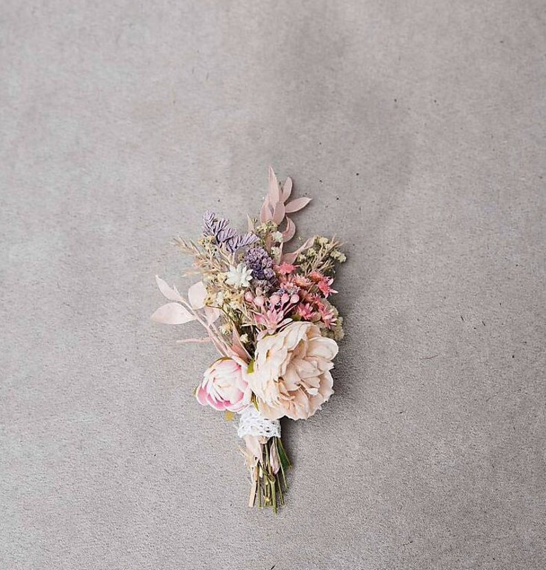 May include: A small, delicate floral arrangement featuring a large, light pink peony and smaller pink, purple, and white flowers. The bouquet is tied with a white lace ribbon and stems. The background is a solid gray.