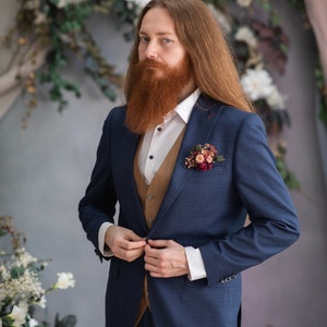 May include: A man with long red hair and a beard is wearing a blue suit with a brown vest. He is adjusting the button on his jacket. He is wearing a white shirt and has a small bouquet of flowers pinned to his lapel.