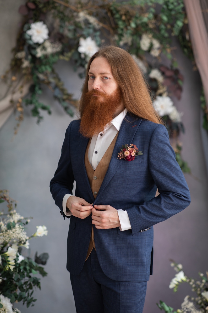 May include: A man with long red hair and a beard is wearing a blue suit with a brown vest. He is adjusting the button on his jacket. He is wearing a white shirt and has a small bouquet of flowers pinned to his lapel.