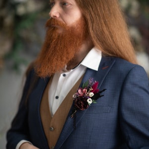 May include: A man wearing a blue suit with a brown waistcoat and a white shirt. He has a long red beard and is wearing a floral buttonhole on his lapel.