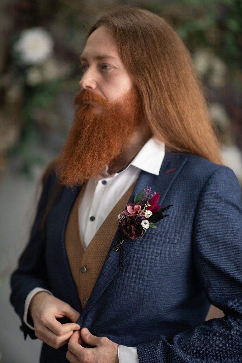 May include: A man wearing a blue suit with a brown waistcoat and a white shirt. He has a long red beard and is wearing a floral buttonhole on his lapel.