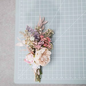 May include: A small, dried flower bouquet featuring a variety of colors. The bouquet includes light pink peonies, lavender, and other dried flowers. The stems are wrapped with a white lace ribbon. The bouquet is on a light blue cutting mat.